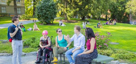 Viisi ihmistä kesäisessä puistossa keskustelemassa. Yksi seisoo, yksi istuu pyörätuolissa ja muut puistonpenkillä.