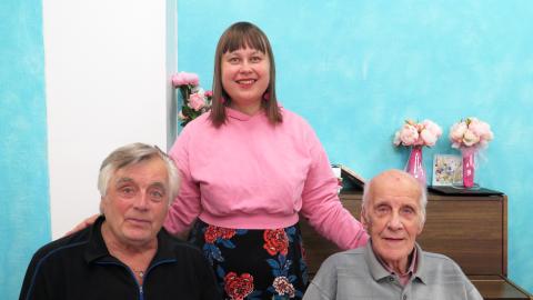 Kaksi iäkästä miestä istuu ja takanaan seisoo vaaleanpunaiseen paitaan pukeutunut nuorempi nainen. Miesten välissä on kukkakimppu.
