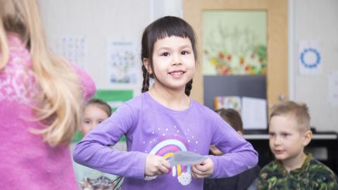 Children in a classroom, a girl with dark hair looks at the camera.