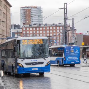 Tampereen keskustassa kaksi sinivalkoista kaupunkiliikenteen bussia toinen menossa ja toinen tulossa. Kadulla ihmisiä bussipysäkillä ja taustalla kaupunkinäkymää.