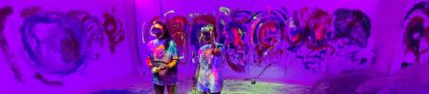 Two performers covered in paint in the middle of purple painted space.