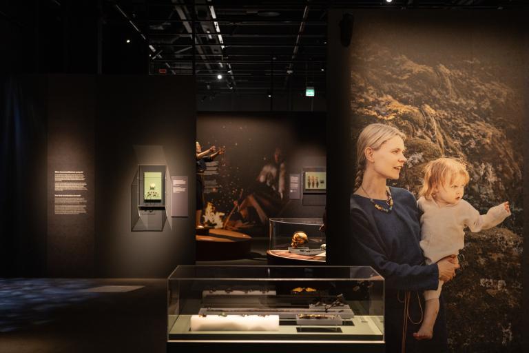 Hämärässä näyttelytilassa esineitä vitriinissä keskellä huonetta. Seinällä kuvassa äiti ja lapsi.