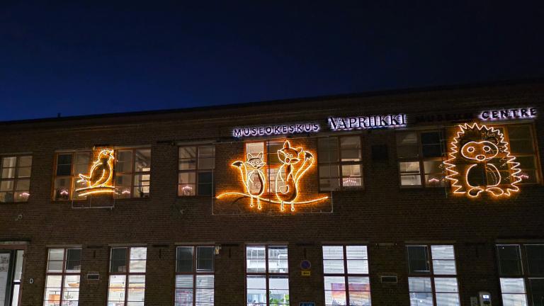 Vapriikin ulkoseinällä eläinvalohahmoja: lintu, kaksi kettua ja siili. Rakennuksen ikkunoista loistaa valoa pimeässä illassa.