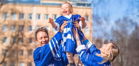Vanhemmat nostavat iloista pientä lasta ylös, kaikki ovat pukeutuneita Tampereen pelipaitoihin.