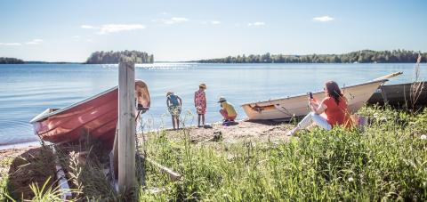 On the shore of a lake, three children explore the sand, a person sits on the edge of the grass with a phone in hand, two boats on docks.