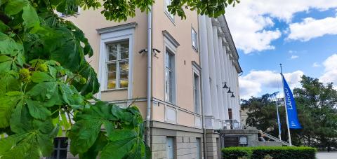 Uusklassinen vaaleankeltainen rakennus kohoaa jyhkeänä maisemassa. Rakennuksen edeessä on pitkä portaikko, jonka sivuilla on siniset banderollit. Rakennusta kehystävät vihreät puiden lehvästöt.