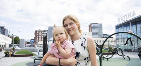 Teini-ikäinen nuori pitelee sylissään parivuotiasta sisarustaan. He istuvat Ratinan kauppakeskuksen lasten leikkipaikan luona.
