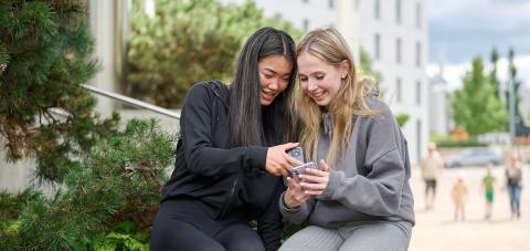 Kaksi nuorta naista istuu Tampere-talon lähellä katsomassa iloisesti puhelinta.