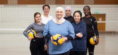 Six women on a volleyball court in a group photo, three of them are holding a volleyball