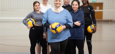Six women on a volleyball court in a group photo, three of them are holding a volleyball