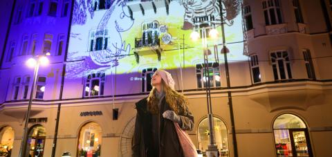 Henkilö seisoo Jugend-torilla ja katsoo ylävasemmalle, taustalla muumiaiheinen valoteos rakennuksen julkisivuun heijastettuna.