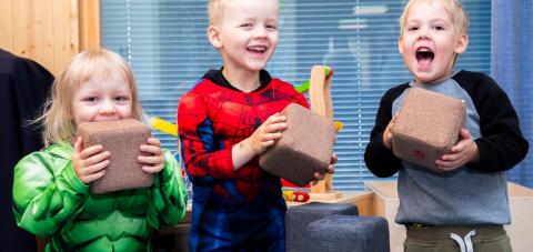 Kolme pientä lasta katsoo kameraan nauraen pitäen pieniä ruskeita pehmokuutioita kädessään.