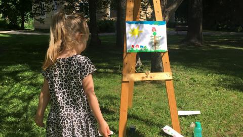 Pieni tyttö seisoo kesäisenä päivänä maalaustelineen edessä. Hän on maalannut auringon ja kukkia.