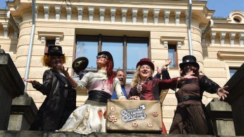 Viisi esiintyjää rooliasuissa vanhan rakennuksen edessä. Kaksi esiintyjistä pitelee vanhaa matkalaukkua, jossa lukee Tampere Steampunk Festival