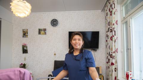 Filippiiniläinen hoiva-avustaja seisoo hymyillen asukkaan huoneessa.