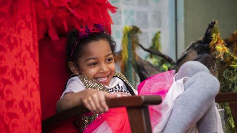 Lapsi istuu punaisessa nojatuolissa roolivaatteet päällä ja nauraa.