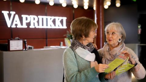 Kaksi iäkästä naista tutkii Vapriikin esitettä Vapriikin aulassa. Taustalla asiakaspalvelutiski.