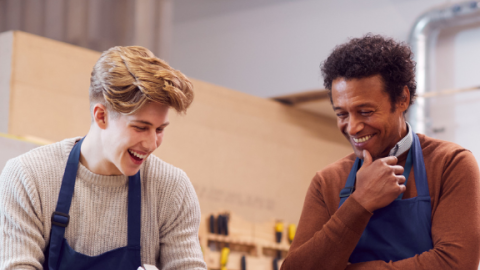 Nuori mies käyttää höylää verstaassa. Hänen vieressään työtä seuraa vanhempi mies. He hymyilevät.