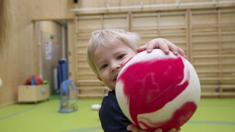 Pieni naurava lapsi pitää isoa punaista palloa käsissään.