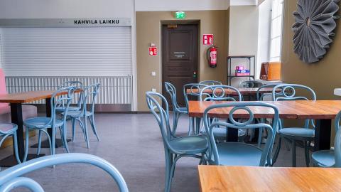 Sinisiä tuoleja ja puupintaisia pöytiä tyhjässä kahvilatilassa. Kahvila on suljettu.