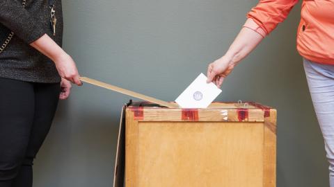 Virkailija avaa äänestyslaatikkoa, ja äänestäjä pudottaa äänestyslipun laatikkoon.