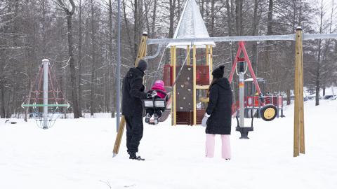 Two adults and a child in a winter playground. The child is swinging on the swing. 