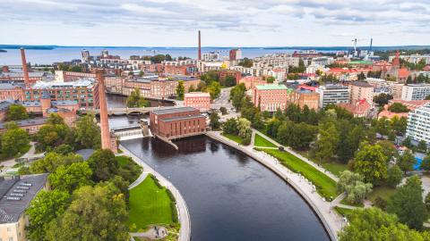Kesäinen Tampereen kaupunkimaisema, jossa näkyy Tammerkoski, tehtaan piippuja, rakennuksia ja takana Näsijärvi.