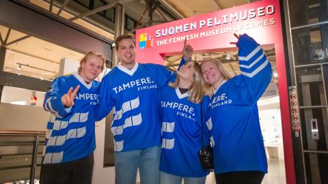 Neljä nuorta henkilöä seisovat Vapriikin Pelimuseon edustalla. Kaikilla on päällä sininen Tampere -kiekkopaita.