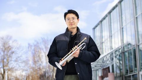 Principal trumpet player Xiang Guo stands in front of the glass wall of Tampere Hall with his trumpet.
