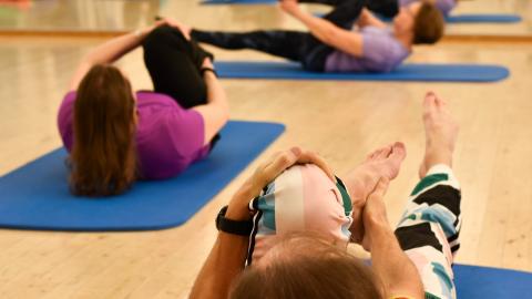 Kolme henkilöä harjoittaa pilatesta lattialla jumppamatoilla maaten. He pitävät toisesta polvestaan kiinni.