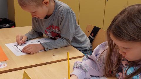 Kaksi lastaa piirtää pulpeteilla. Toisella on suu mutrussa keskittymisestä.