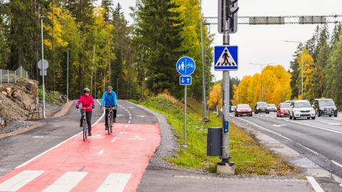 Kaksi pyöräilijää pyörätiellä ja autoliikennettä kadulla syksyllä.