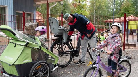 Hilla Lyly, Pihla Lyly and Jani Lyly in the kindergarten yard with two bicycles, one of which has a trailer.