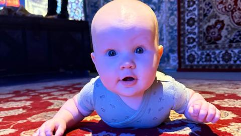 Vauva mahallaan lattialla punaisen maton päällä, katsoo suoraan kameraan.