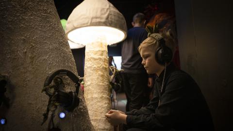 Lapsi istuu näyttelytilassa suurten valaistujen sienien keskellä kuulokkeet korvilla.