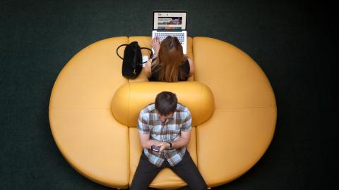 Ylhäältä päin kuvattuna kaksi henkilöä istuu ja käyttävät toinen kannettavaa tietokonetta ja toinen älypuhelinta.