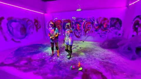 Violettisävyisessä kuvassa kaksi henkilöä virtuaalilasit päässä huoneessa, jossa taidetta seinillä.