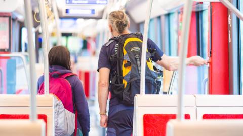 Kaksi henkilöä seisoo ratikkavaunun käytävällä selin. Etualalla on istuimia ja taustalla reittitaulu.