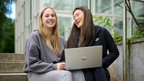 Kaksi nuorta naista istuvat yliopistokampuksen ulkorappusilla tietokoneen kanssa ja nauravat.