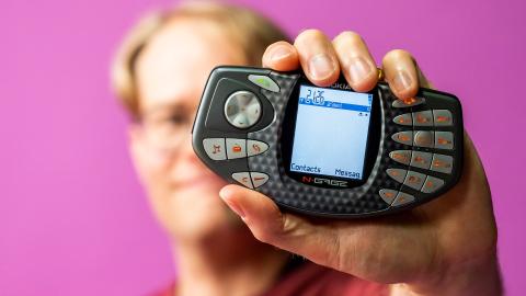 Mies pitelee edessään kädessä pelilaitetta, jossa on keskellä näyttö ja molemmilla sivuilla näppäimiä. Tausta on vaaleanpunainen.