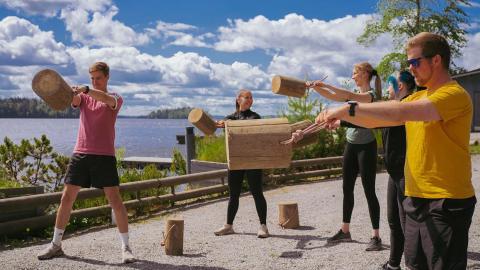 Viisi ihmistä kuntoilee köyden päässä olevia pöllejä heilutellen kesäisen järven rannalla.