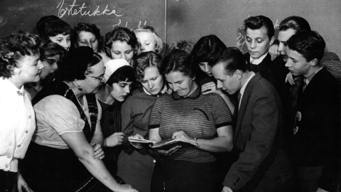 Joukko opiskelijoita on kokoontunut opettajan ympärille tutkimaan kirjaa.