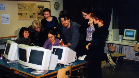 Seitsemän henkilöä seisoo tietokoneiden ympärillä luokkaympäristössä.