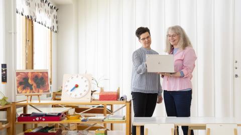 Two women looking at a laptop. On their left a shelf with miscellanous items, including children&#039;s toy clock.
