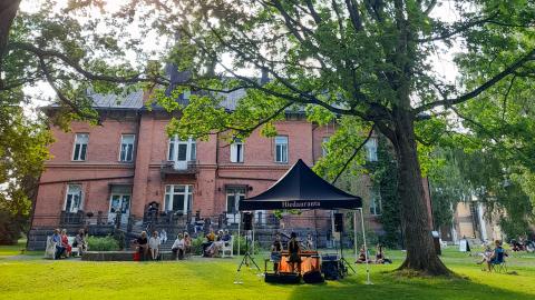 Kesäiseen puistoon on kokoontunut ihmisiä kuuntelemaan konserttia. Esiintyjä soittaa kosketinsoitinta telttakatoksen alla.