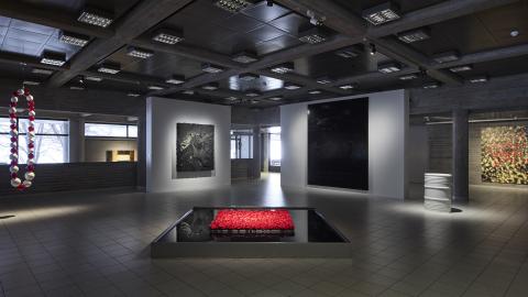 View from the ground floor of the museum, dimly lit. Huge red and white glass beads hang from the ceiling, behind which is a landscape window looking out onto a snow-covered sculpture park. In the centre of the picture is a large pool of ink with red roses in the middle. Behind it are two white partitions, one with a black non-representational relief hanging on it, the other with a black painting almost as high as the partition with representational elements. Both the relief and the painting are reflected i