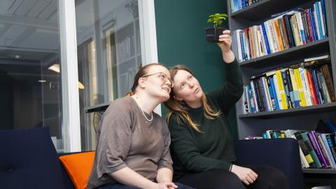 Two women sitting on the couch. The other one holds a small plant in a pot, which both are looking at. On the right there&#039;s a book shelf.