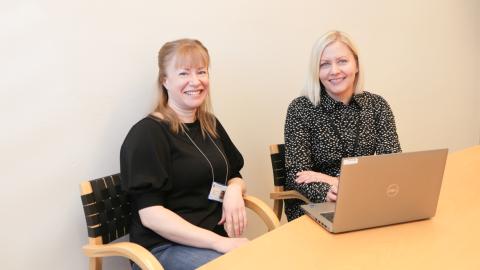 Kaksi henkilöä istuu pöydän ääressä tietokoneen kanssa ja katsovat hymyillen kameraan.