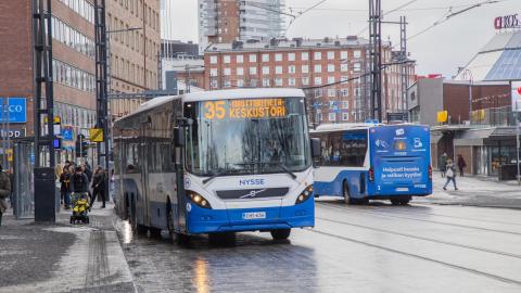 Tampereen keskustassa kaksi sinivalkoista kaupunkiliikenteen bussia toinen menossa ja toinen tulossa. Kadulla ihmisiä bussipysäkillä ja taustalla kaupunkinäkymää.