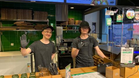 Kaksi PizzaCastlen työntekijää seisovat ravintolan tiskin takana ja vilkuttavat.